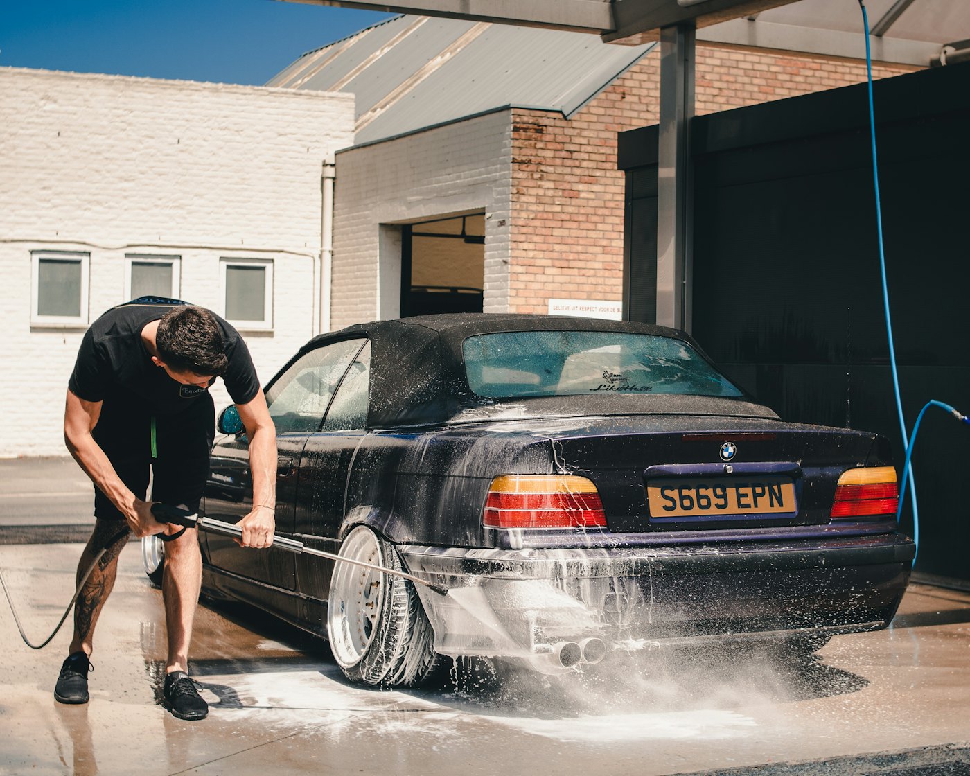 A DnA detailer rinsing a navy sedan in the driveway of a brick Eastside home